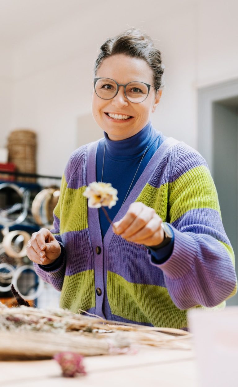 Inhaberin IMMERBLUME Studio Nadin Hoppe im Atelier Frau namens Nadin Hoppe mit getrockneten Amaranth auf dem Arm, trägt einen roten Pullover und blaue Hose, lächelt.