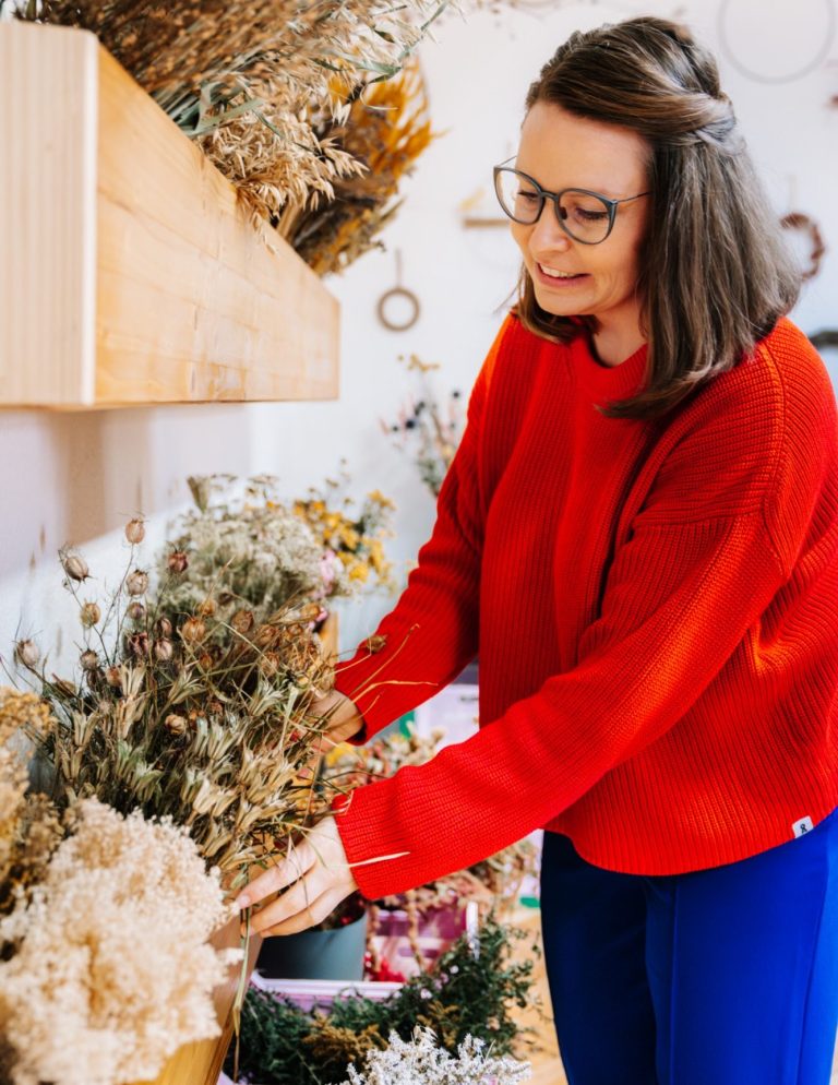 Frau in rotem Pullover arrangiert Trockenblumen an einem Regal voller Trockenblumen